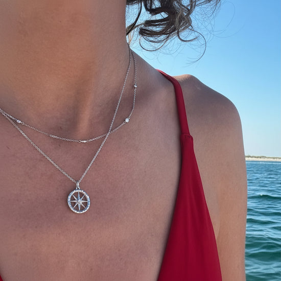 Person wearing a gold compass necklace with a pendant by the water